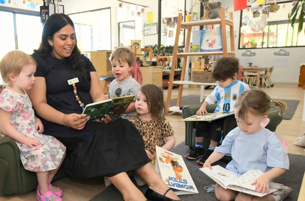 An educator sitting on a low armchair reading a picture book with a group of toddlers, while two other children look through their own books